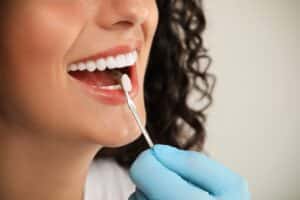 Close-up of a dentist evaluating a young woman's tooth color for dental veneer treatment against a light grey background